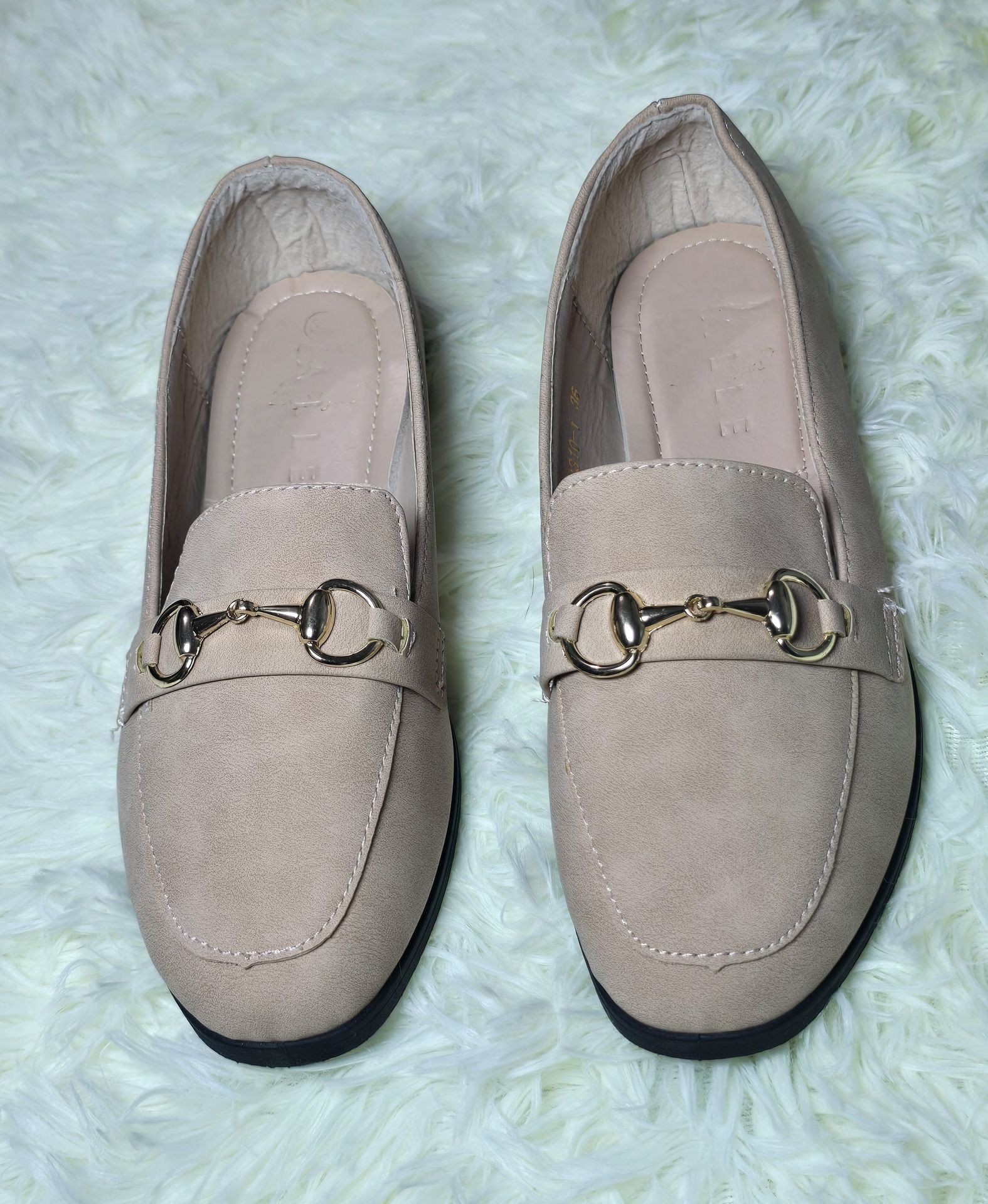 Pair of beige loafers with gold buckle detailing, placed on a light green shaggy rug.