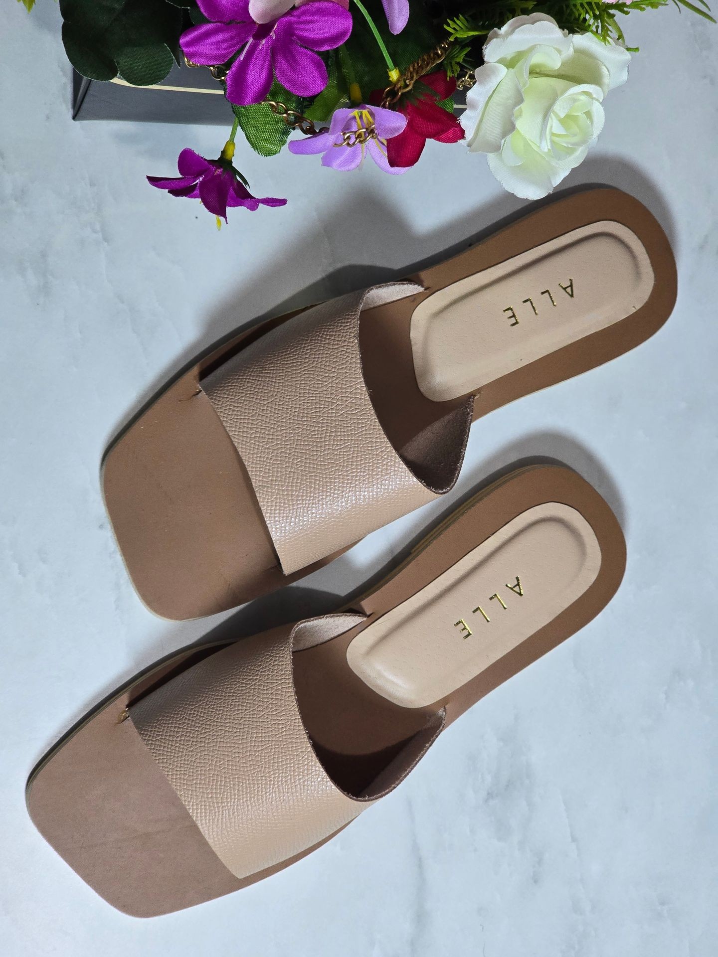 Pair of beige slide sandals on a marble surface, accompanied by a bouquet of artificial flowers.