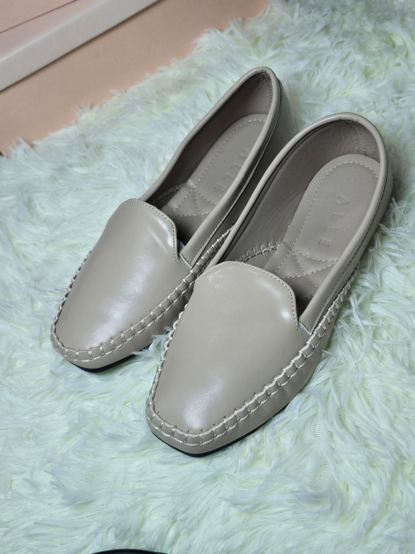 Pair of beige leather loafers with stitched detailing on a light green fluffy rug.