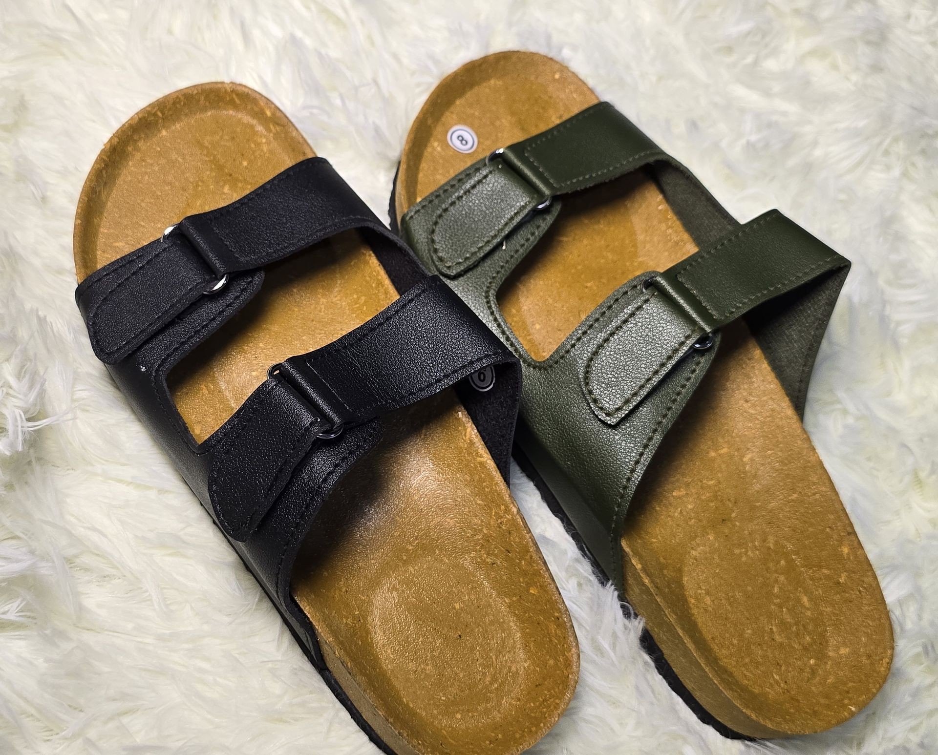 Black and green adjustable strap sandals with cork footbeds on a white, fluffy surface.