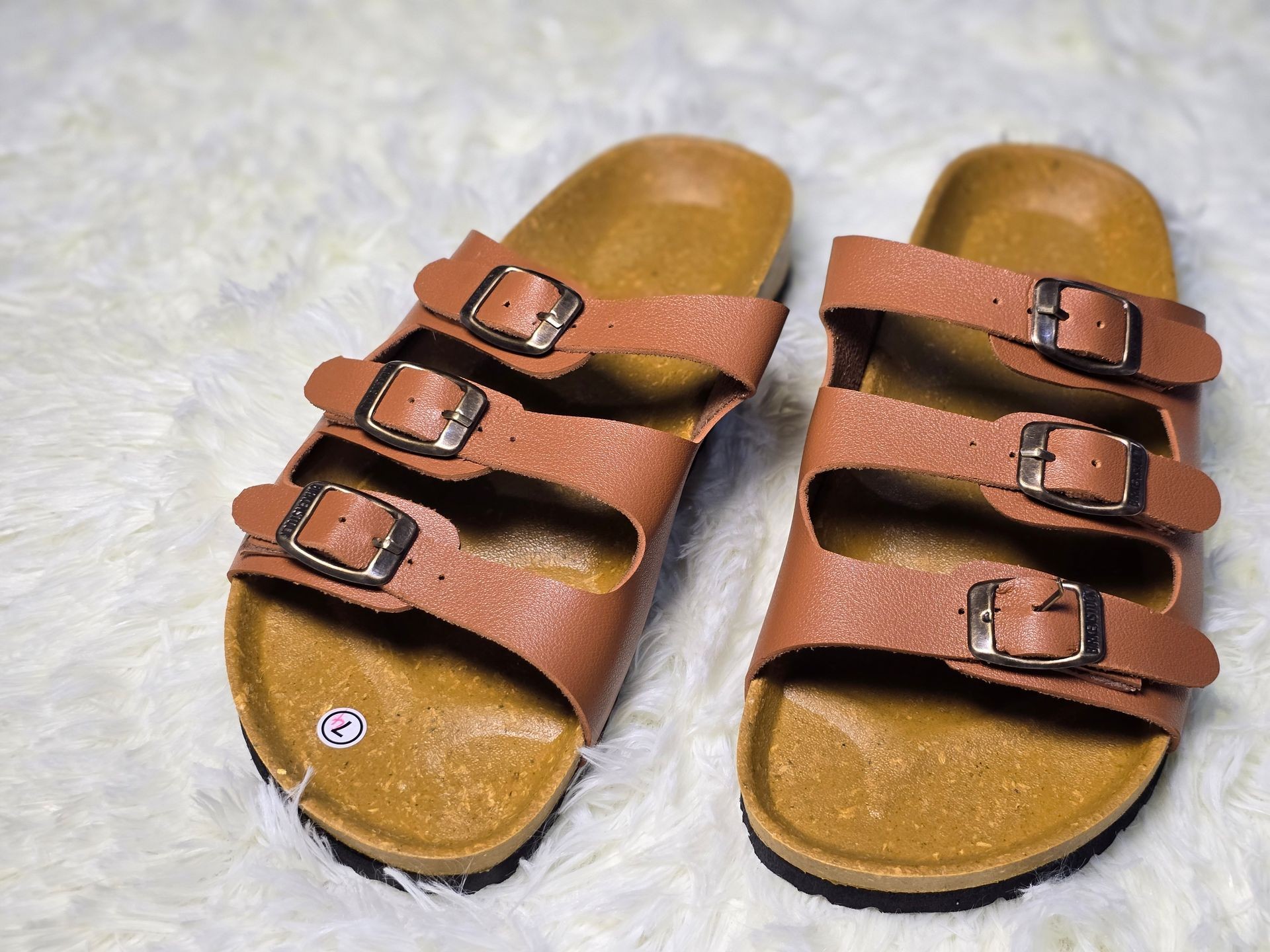 Close-up of tan sandals with three adjustable buckle straps, resting on a fluffy white surface.