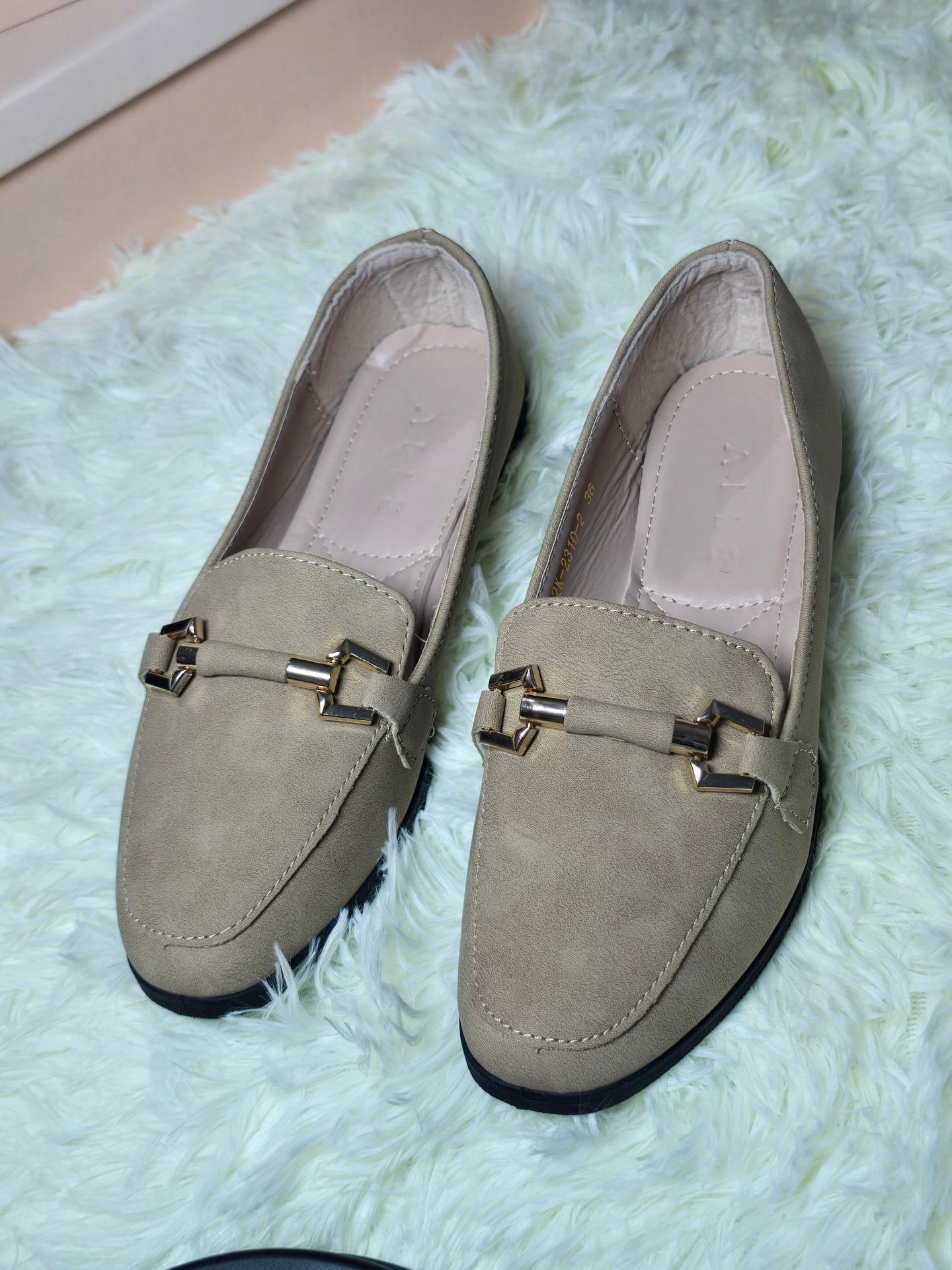 Pair of beige loafers with gold buckles placed on a fluffy white rug.