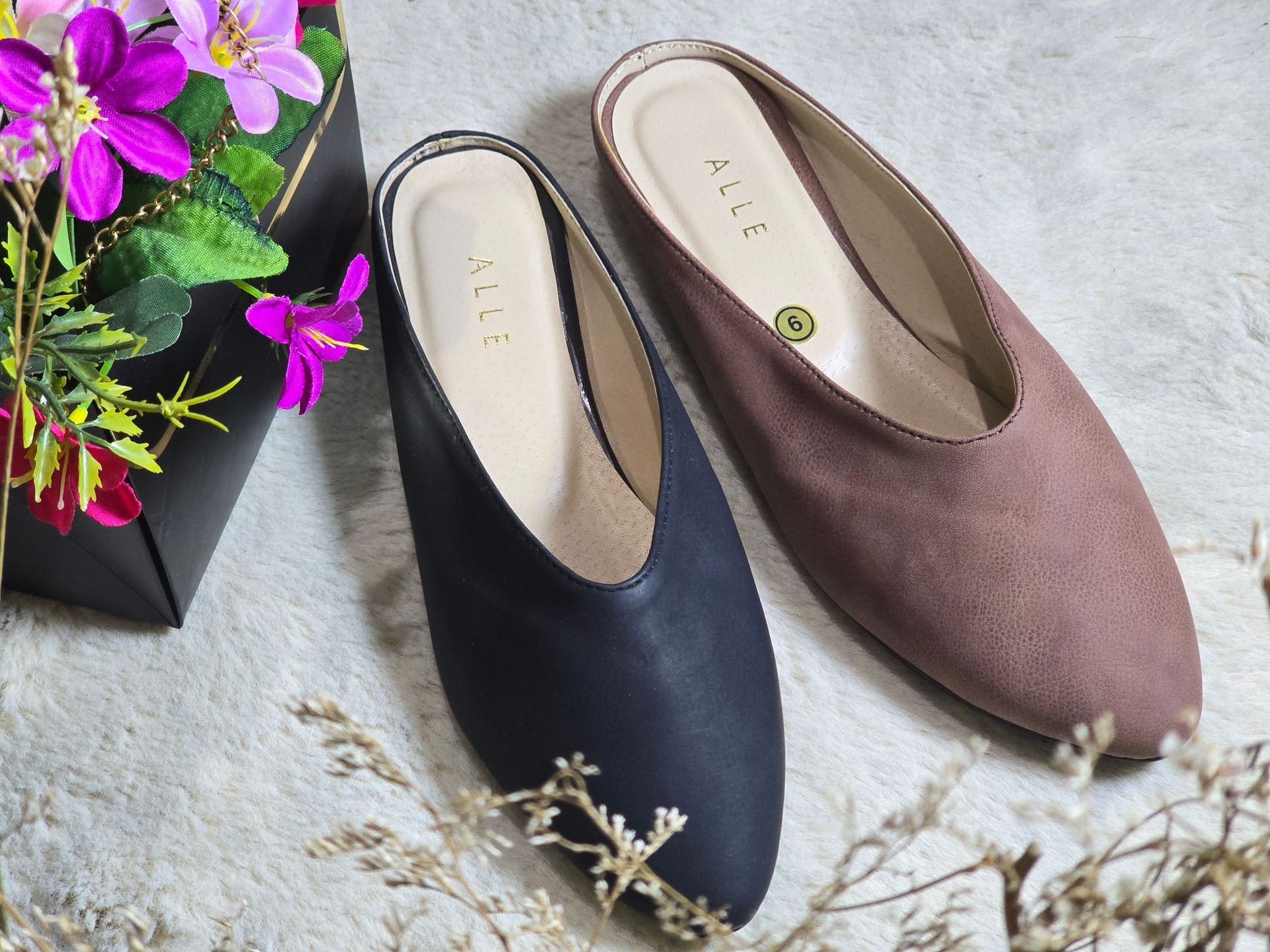 Pair of women's slip-on shoes in black and brown on a soft surface with decorative flowers nearby.