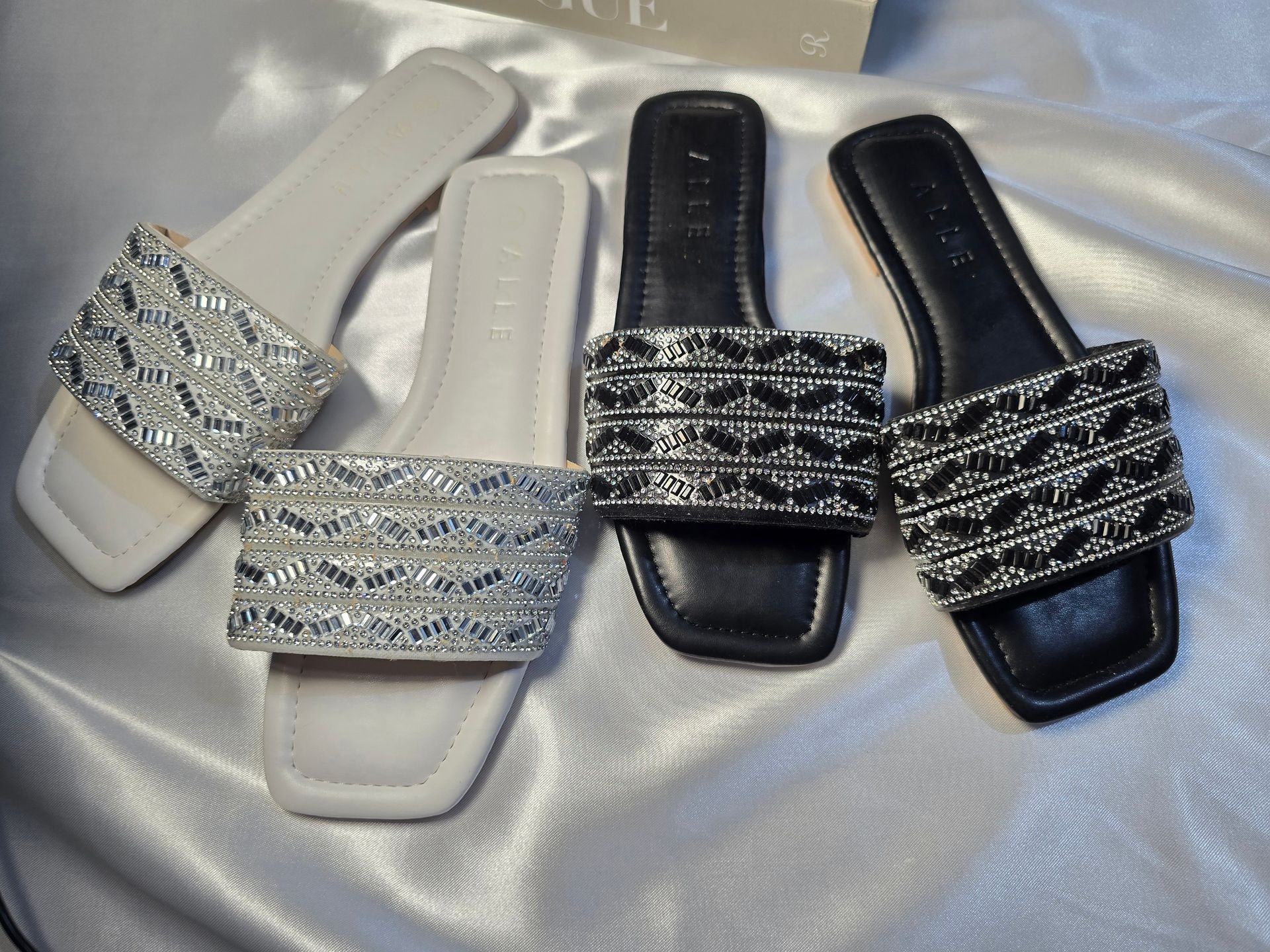 Two pairs of embellished sandals, one white and one black, placed on a silky fabric background.