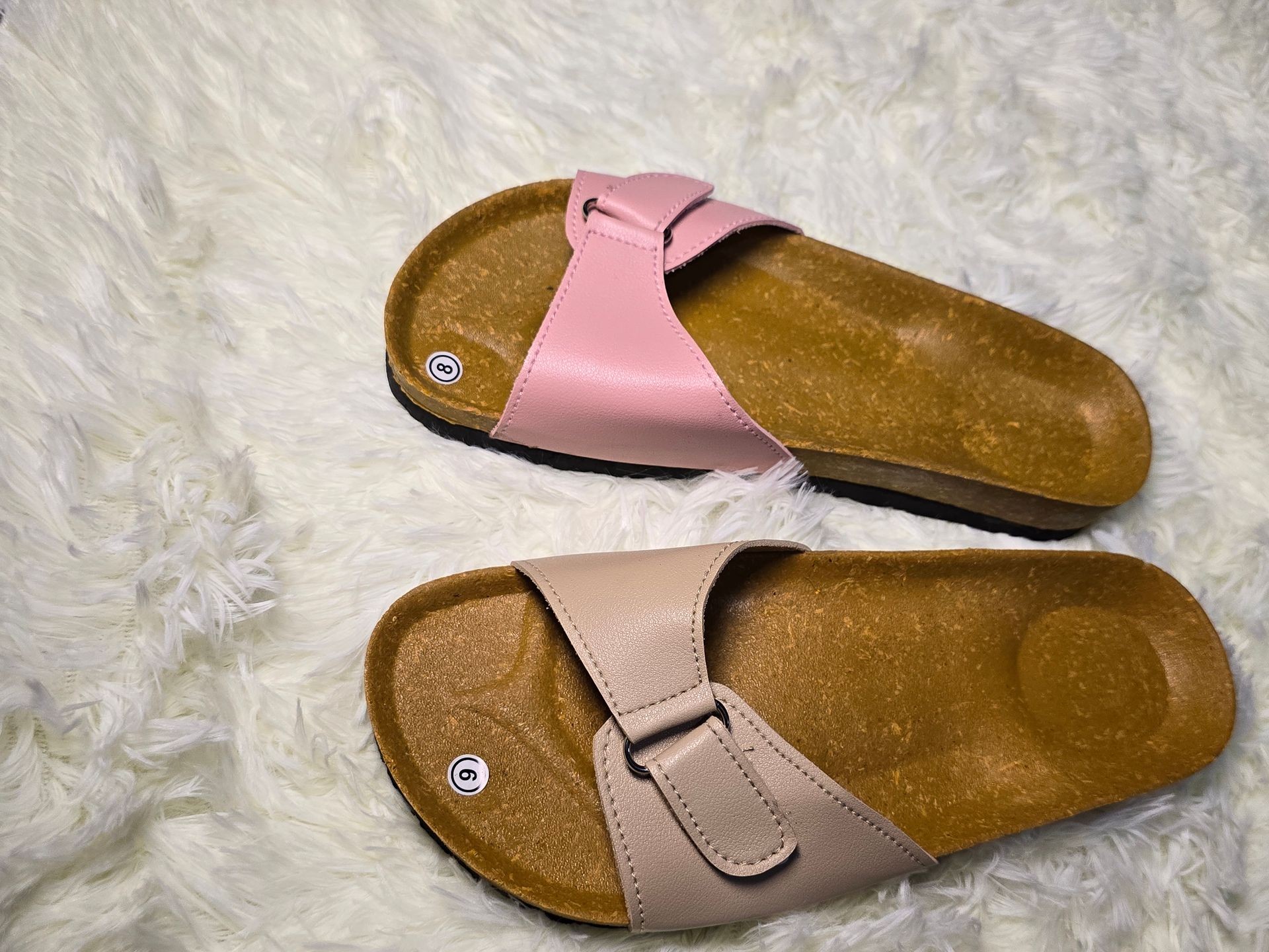 Two pairs of sandals, one pink and one beige, resting on a fluffy white surface.